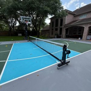 A custom multi sport court in Friendswood, Texas featuring a 50’x30’ half-court with a high school 3-point basketball line, an in-ground Ryval hoop, and a roll-away pickleball net.