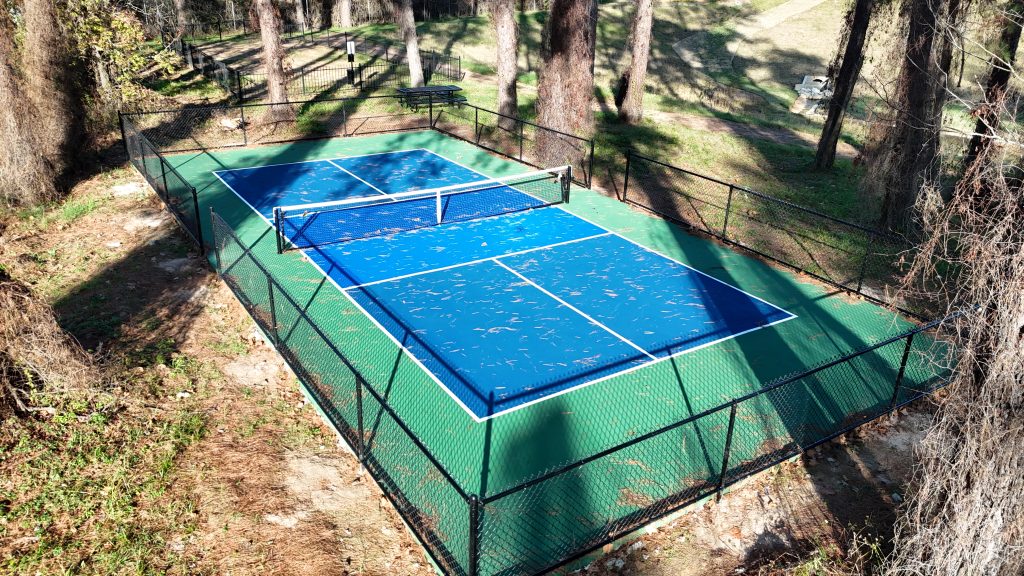 Ground view of fenced blue pickleball court surfacing in Spring Texas wooded area. Title: Fenced Blue Pickleball Court Ground View Spring TX.