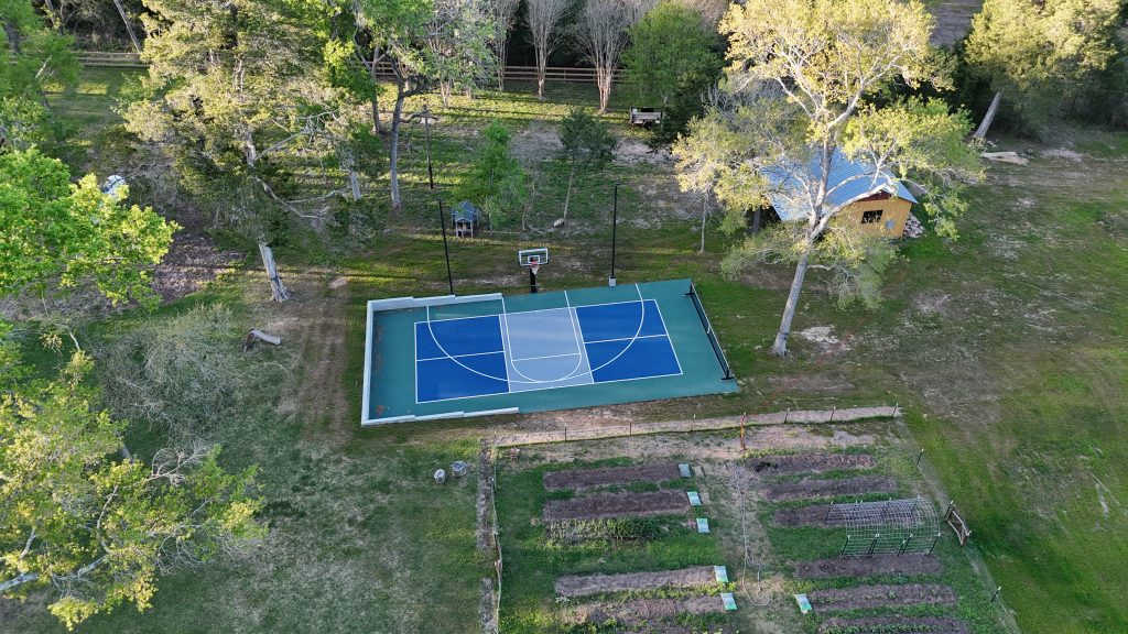 Outdoor basketball court with blue playing surface and hoop in a large residential backyard